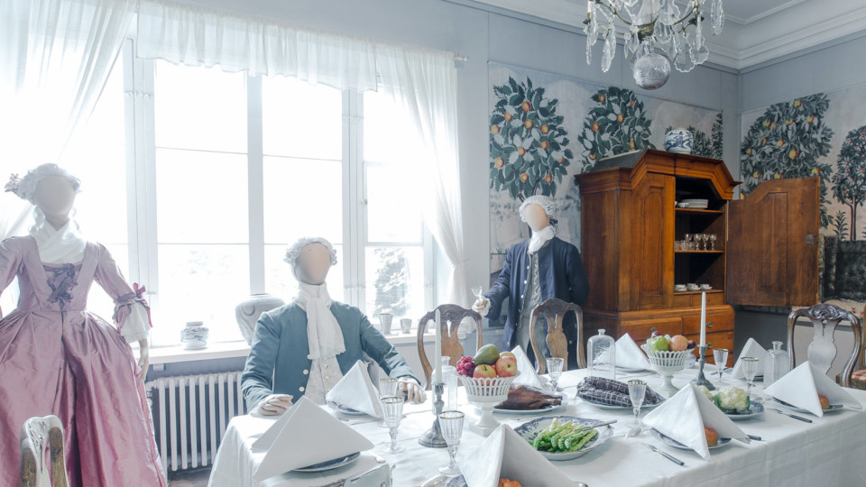 Ruokasali kattauksineen, 1700-luvun aateliston asuihin puetut mallinuket pöydän ääressä.