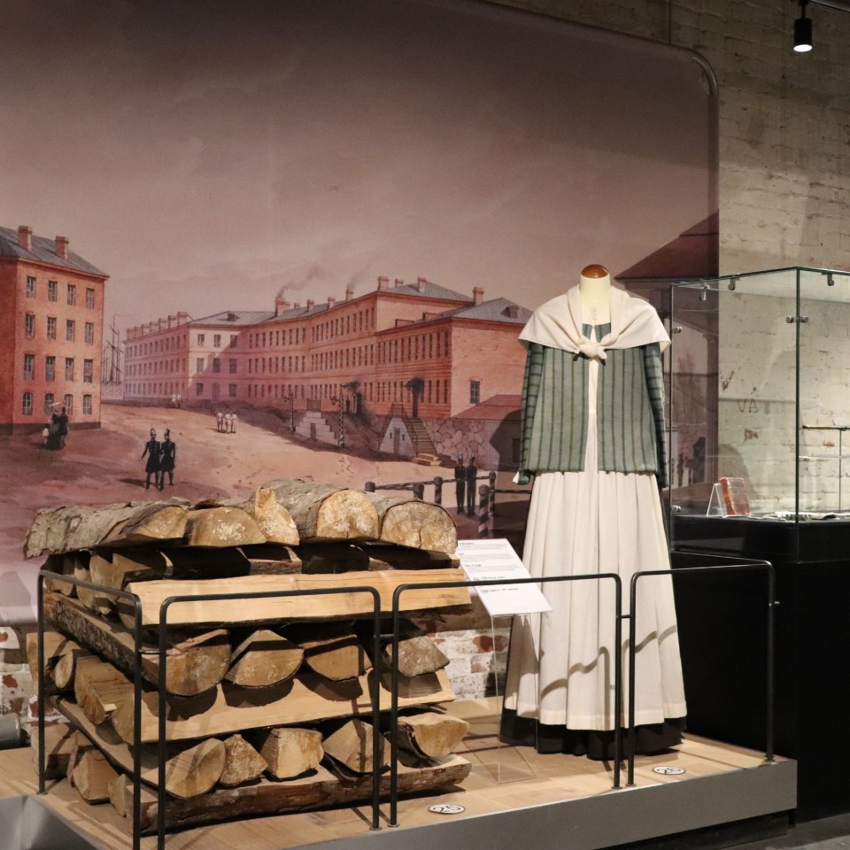 Museossa esillä oleva halkopino sekä 1700-luvun naisen asu. Taustalla maalaus Iso Mustasaaren asuinrakennuksista ja niiden keskellä olevasta aukiosta.