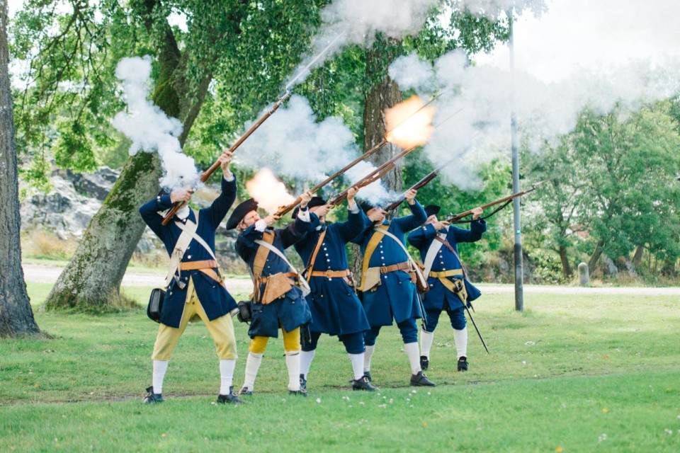 1700-luvun aateliston tyyliin pukeutuneet herrat ampuvat ilmaan kivääreillä.
