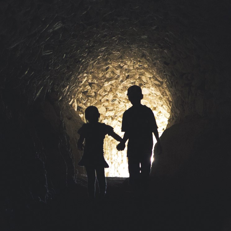 Pimeä tunneli, jonka päässä loistaa valo luoden kahden käsi kädessä kulkevan lapsen siluetin.