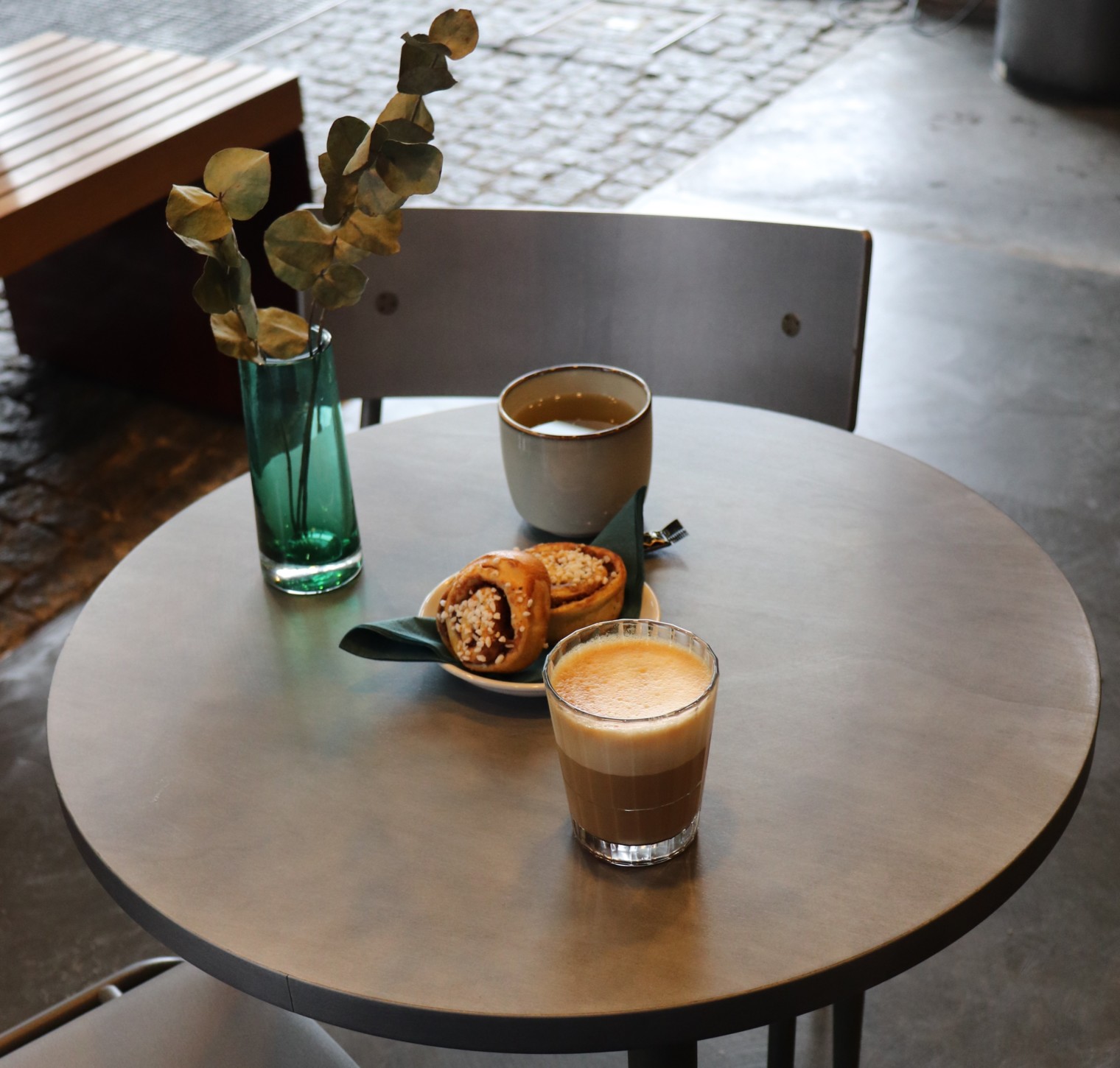 Pyöreä pöytä, jolle katettu kanelipullaa sekä kuppi kahvia ja teetä, pöytää koristaa lasimalja, jossa eukalyptuksen oksia.