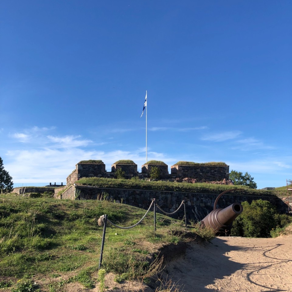 Kustaanmiekan hiekkapolku, joka johtaa tykille. Taustalla kohoaa linnoituksen muurit ja suomenlippu joka liehuu sinitaivasta vasten.